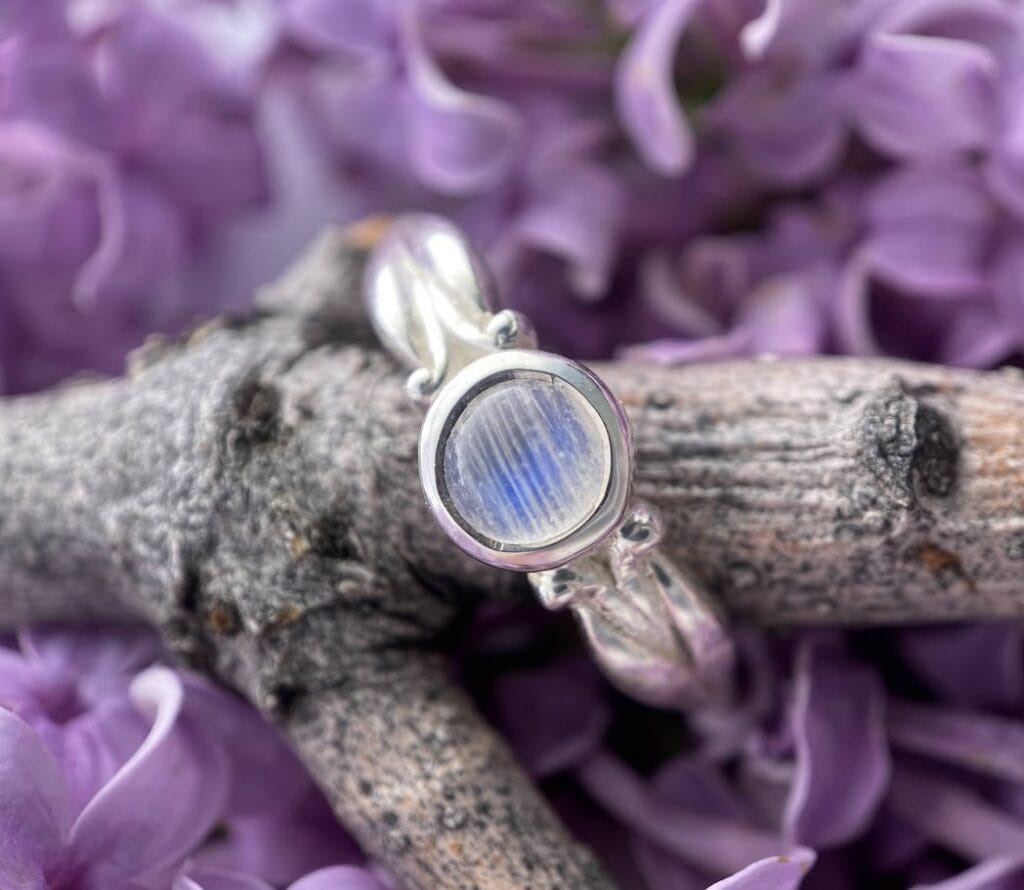 a small circular white moonstone and sterling silver ring laid on a branch against lilac flowers